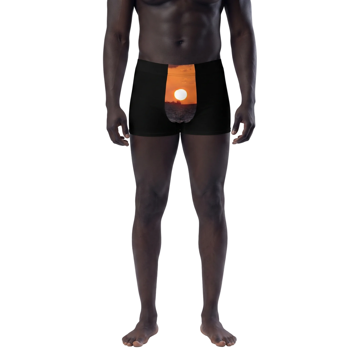 A male model wearing black boxer briefs with an orange and white circular graphic on the front, shown against a plain white background. A small inset at the lower left shows a hand holding a white rectangular item.