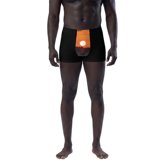 A male model wearing black boxer briefs with an orange and white circular graphic on the front, shown against a plain white background. A small inset at the lower left shows a hand holding a white rectangular item.