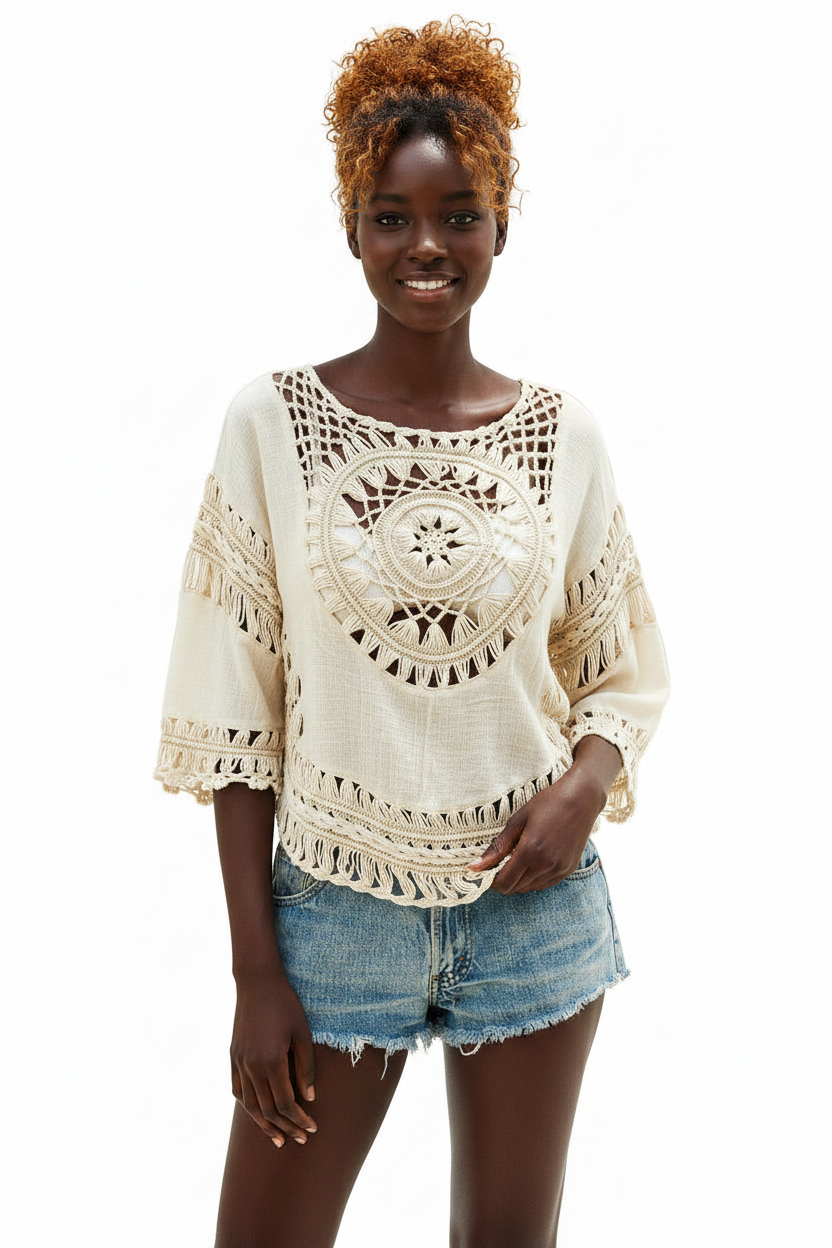 A woman modeling a cream-colored crochet knit top with bohemian-style cutouts and fringed details, paired with denim shorts, against a plain white background.
