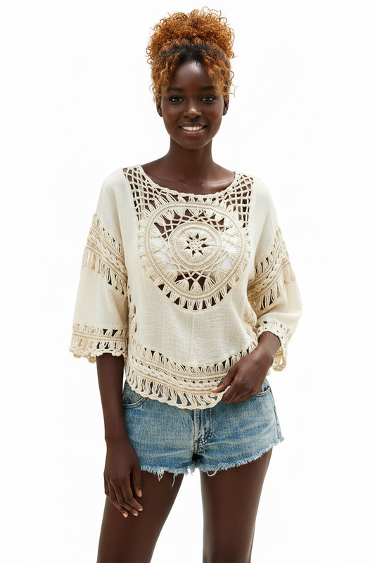 A woman modeling a cream-colored crochet knit top with bohemian-style cutouts and fringed details, paired with denim shorts, against a plain white background.