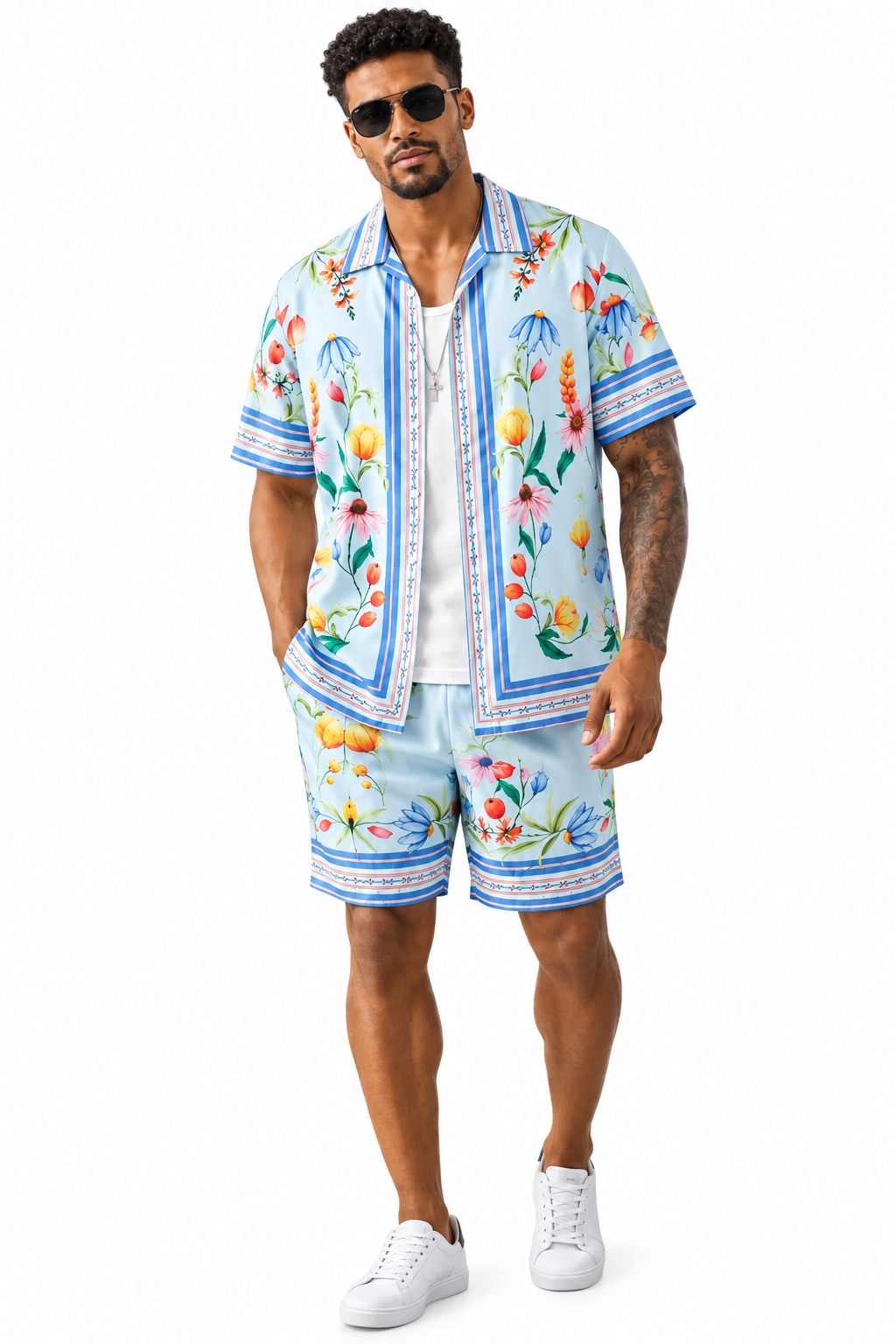 A man modeling a matching light blue short-sleeve button-up shirt and shorts set with a colorful tropical fruit print, styled with white sneakers and sunglasses against a plain white background.
