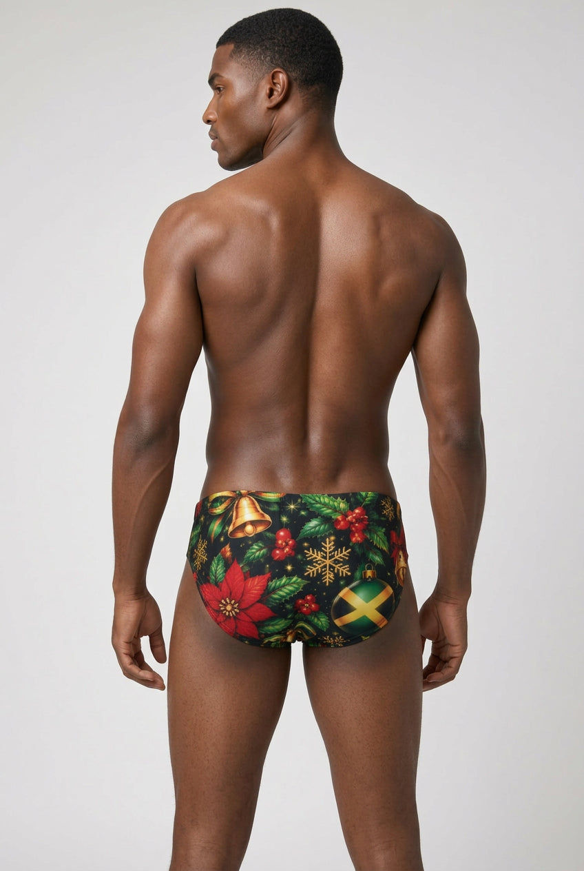 A man shown from the back wearing low-rise swim briefs with a festive Christmas print featuring red poinsettias, green holly leaves, gold bells and snowflakes, and a Jamaican flag motif, against a light gray studio background.