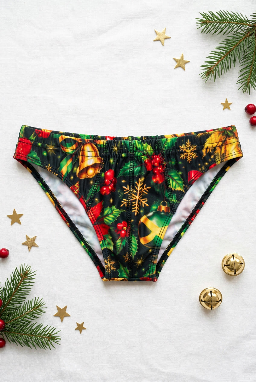 A pair of low-rise bikini bottoms with a festive Christmas print, displayed on a cream-colored mannequin torso. The print features green holly leaves, red berries, gold bells, and gold snowflakes on a dark background.