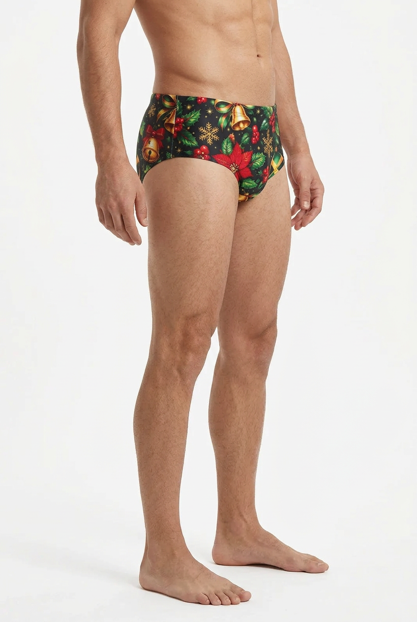 A man modeling a pair of men's briefs with a festive Christmas print featuring red poinsettias, green leaves, gold bells, and pinecones on a dark background, photographed against a clean white studio backdrop.